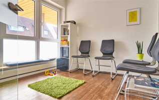 Wartezimmer mit vier grauen Stühlen, einem kleinen Tisch mit Zeitschriften, einem grünen Teppich, einem Regal mit Büchern und einer Pflanze in einem gelben Topf.